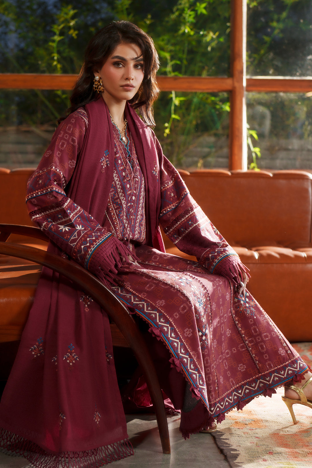 A woman models a traditional Pakistani salwar kameez suit in a warm indoor setting. The outfit is a deep maroon/burgundy color with intricate multicolor embroidery and geometric border patterns.