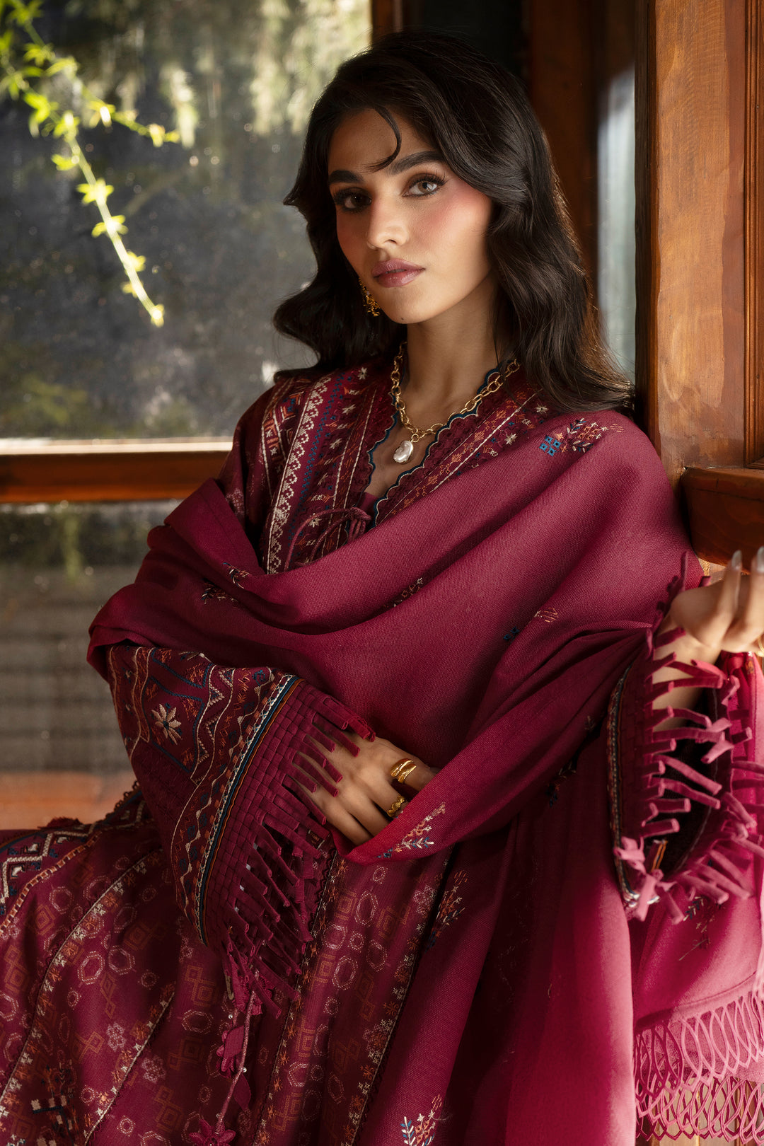 A woman wearing a deep magenta-pink traditional outfit with intricate gold embroidery and a matching fringed shawl, posed in front of a warm wooden window.