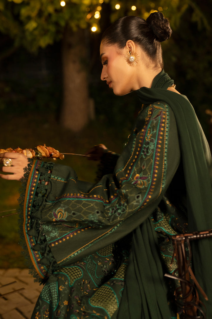A woman in profile wearing a dark green embroidered traditional outfit with intricate patterns and a matching draped scarf, photographed outdoors at night with warm string lights in the background.