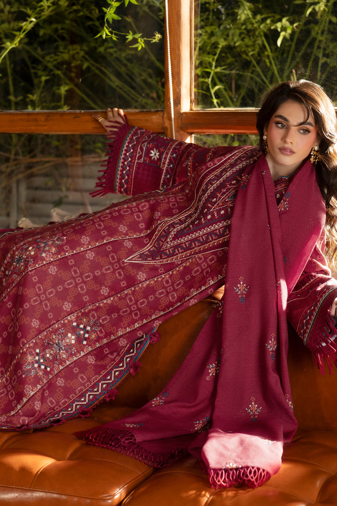 A woman reclines on a brown leather sofa wearing a deep magenta embroidered traditional outfit with a matching scarf, set in front of a window with green foliage visible outside.