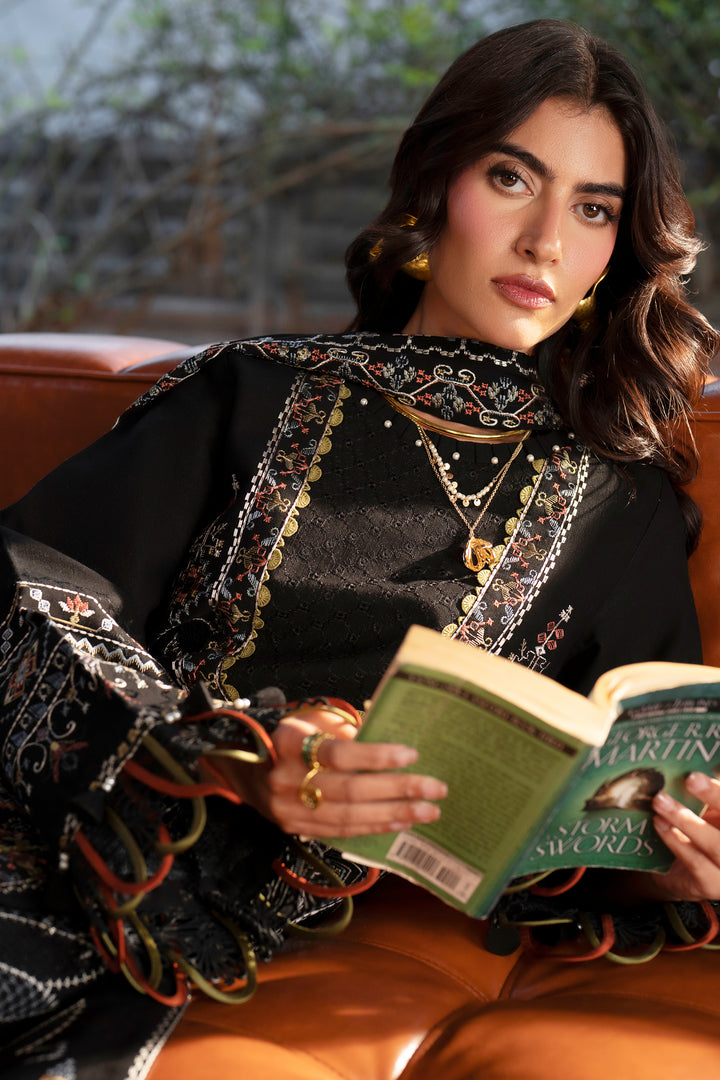 A woman wearing a black embroidered traditional outfit with gold jewelry sits on a brown leather couch, holding an open book.