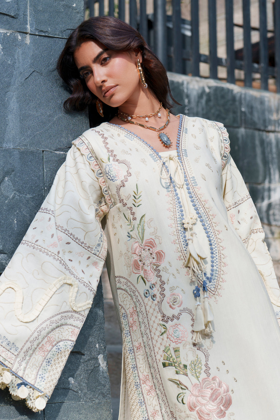 A woman wearing a cream-colored embroidered kurta with floral patterns and tassel ties, posing against a dark gray stone wall.