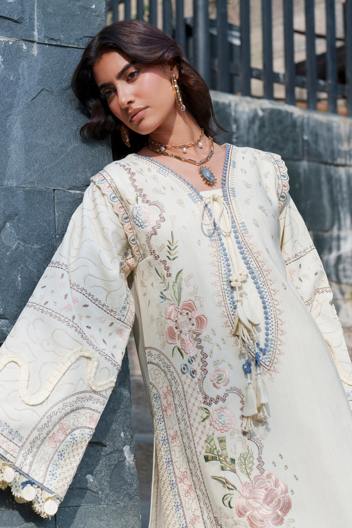 A woman wearing a cream-colored embroidered kurta with floral patterns and tassel ties, posing against a dark gray stone wall.