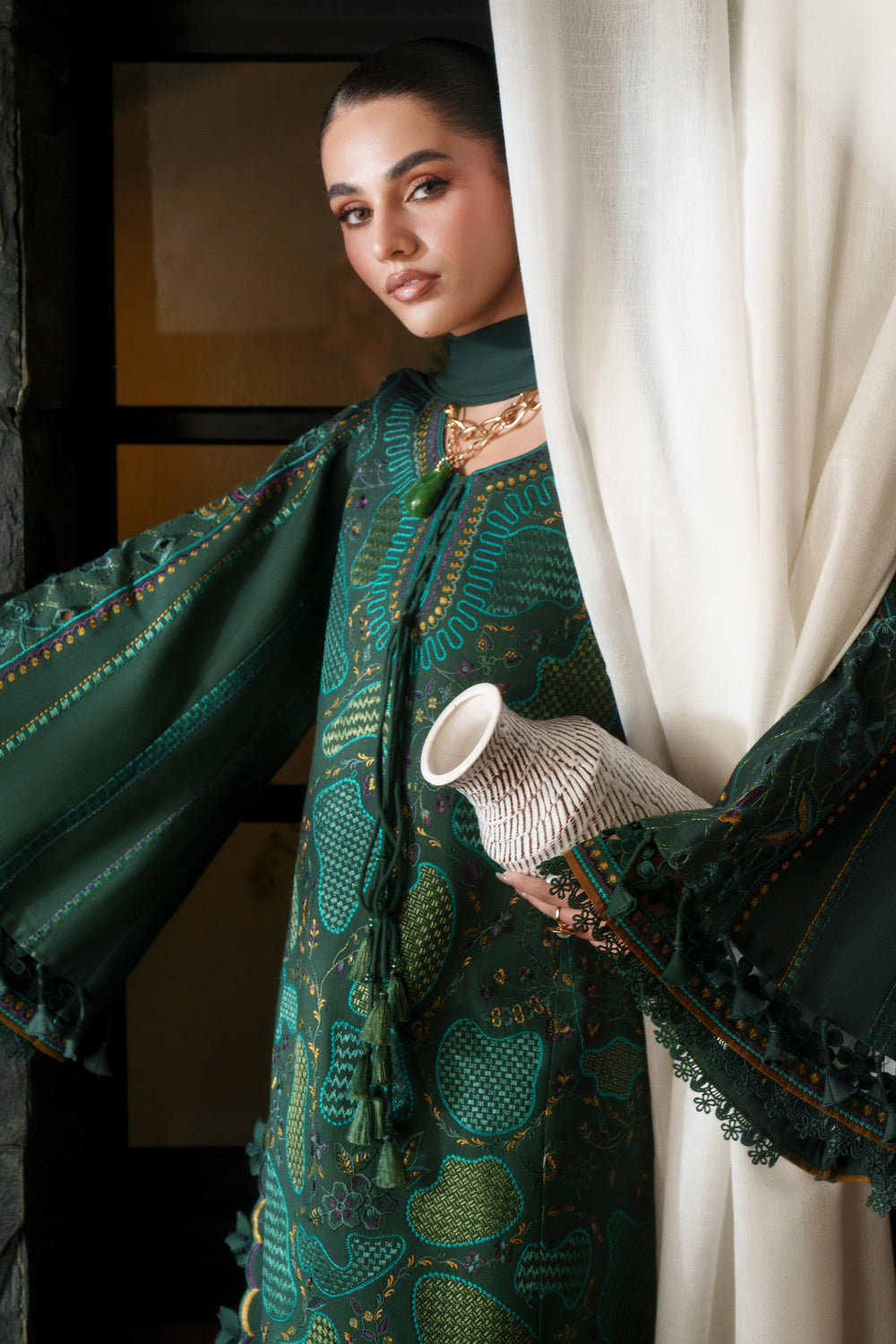 A woman models a dark green embroidered traditional outfit with wide sleeves and a tassel detail, holding a white woven cup in front of a white curtain.